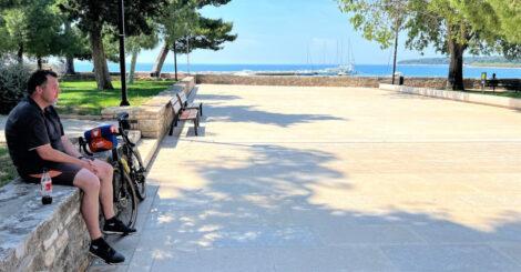 Ein Mann sitzt unter Kieferbäumen auf einer kleinen Steinmauer eines Parks vor einer großen Freifläche neben einem an die Mauer gelehnten Fahrrad. Im Hintergrund ist das blaue Meer zu sehen.