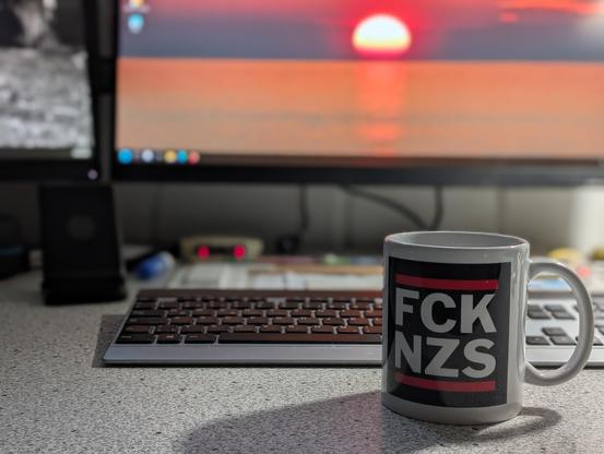 Eine Kaffeetasse mit der Aufschrift "FCK NZS" cir einer Tastatur und zwei Monitoren.