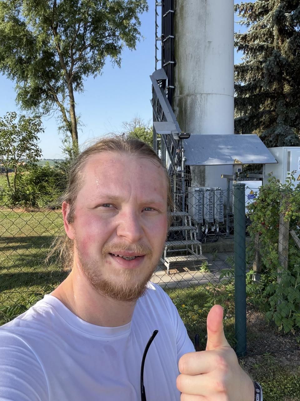 Ein verschwitzter Läufer zeigt mit erhobenem Daumen in die Kamera und steht vor der technischen Anlage eines Mobilfunkmasts. Die Aufnahme vermittelt Erschöpfung und Zufriedenheit nach einer intensiven Laufeinheit an einem sonnigen Sommertag.