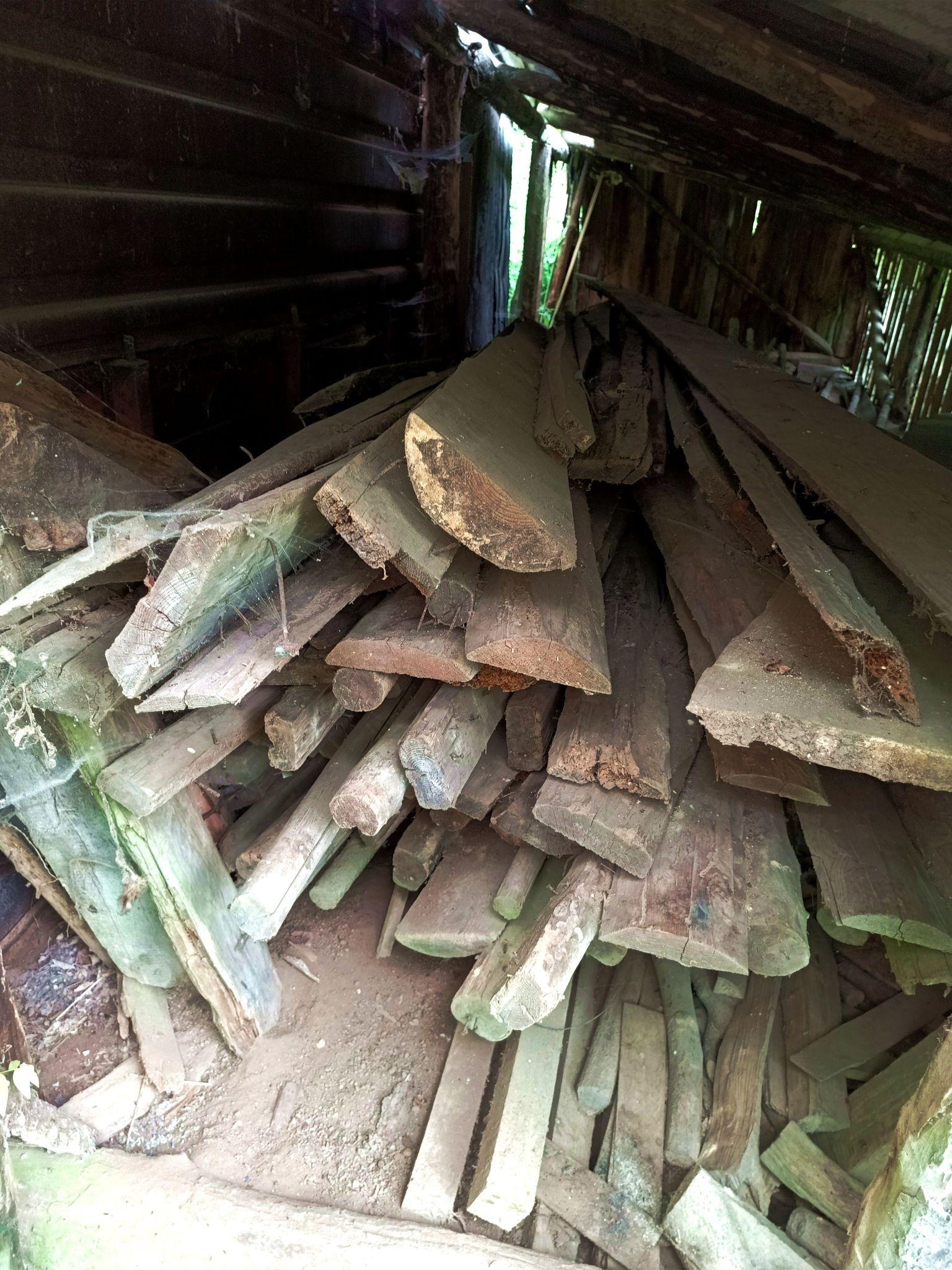 A view into the dark lean-to, with some light streaming in through an opening in the back. The space is half filled with various long lengths of timber, some single cut sides and some beams and some round poles. Everything is dusty and many of them have old nails in them.