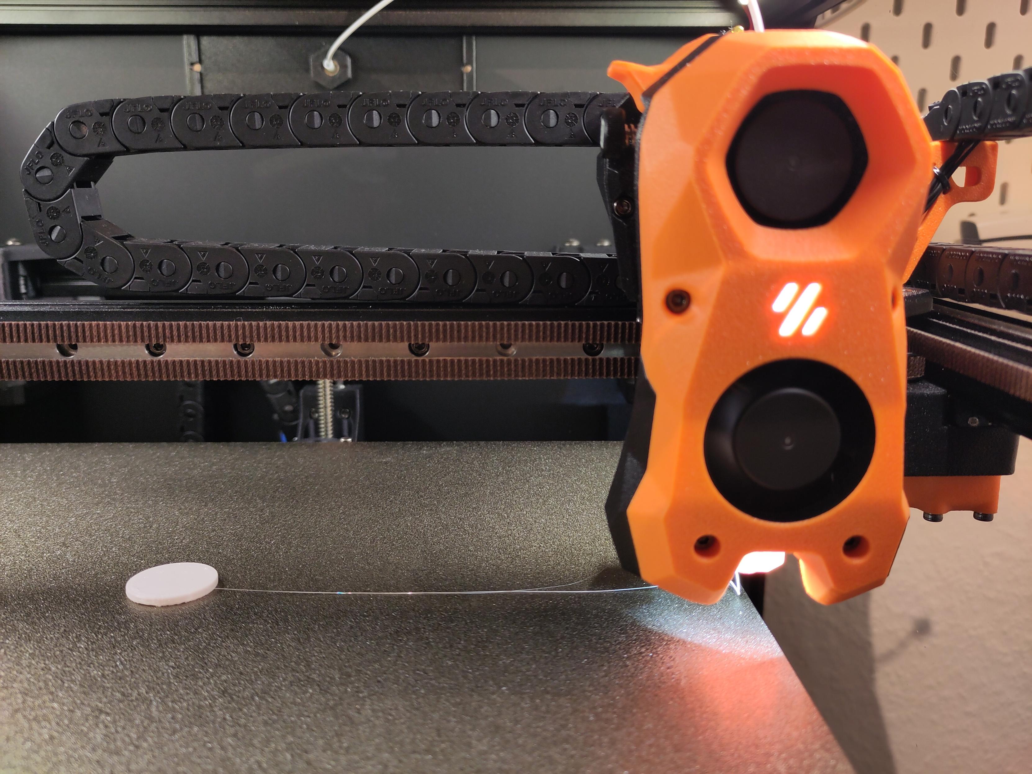 First successful print from a freshly built Voron Trident, a small coin-like cylinder printed from whilte PLA, in the center of the print bed. The toolhead is illuminated from the built-in LED and is parked to the side.