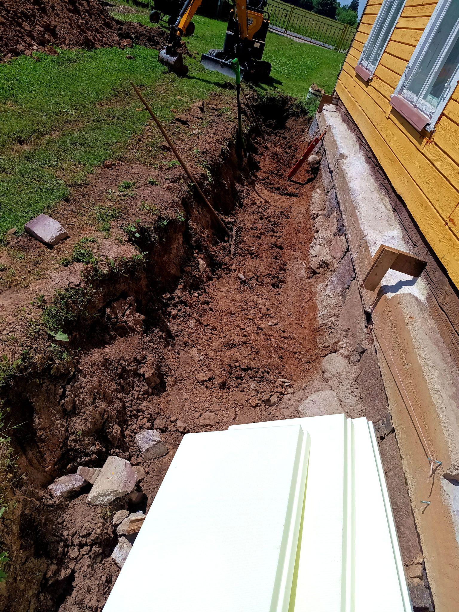 Trench next to an old house, now leveled and with a lot of dirt and rocks adjacent to the old foundation stones removed. Three XPS foam panels are laying in the trench. A mini-excavator is parked at the far end.