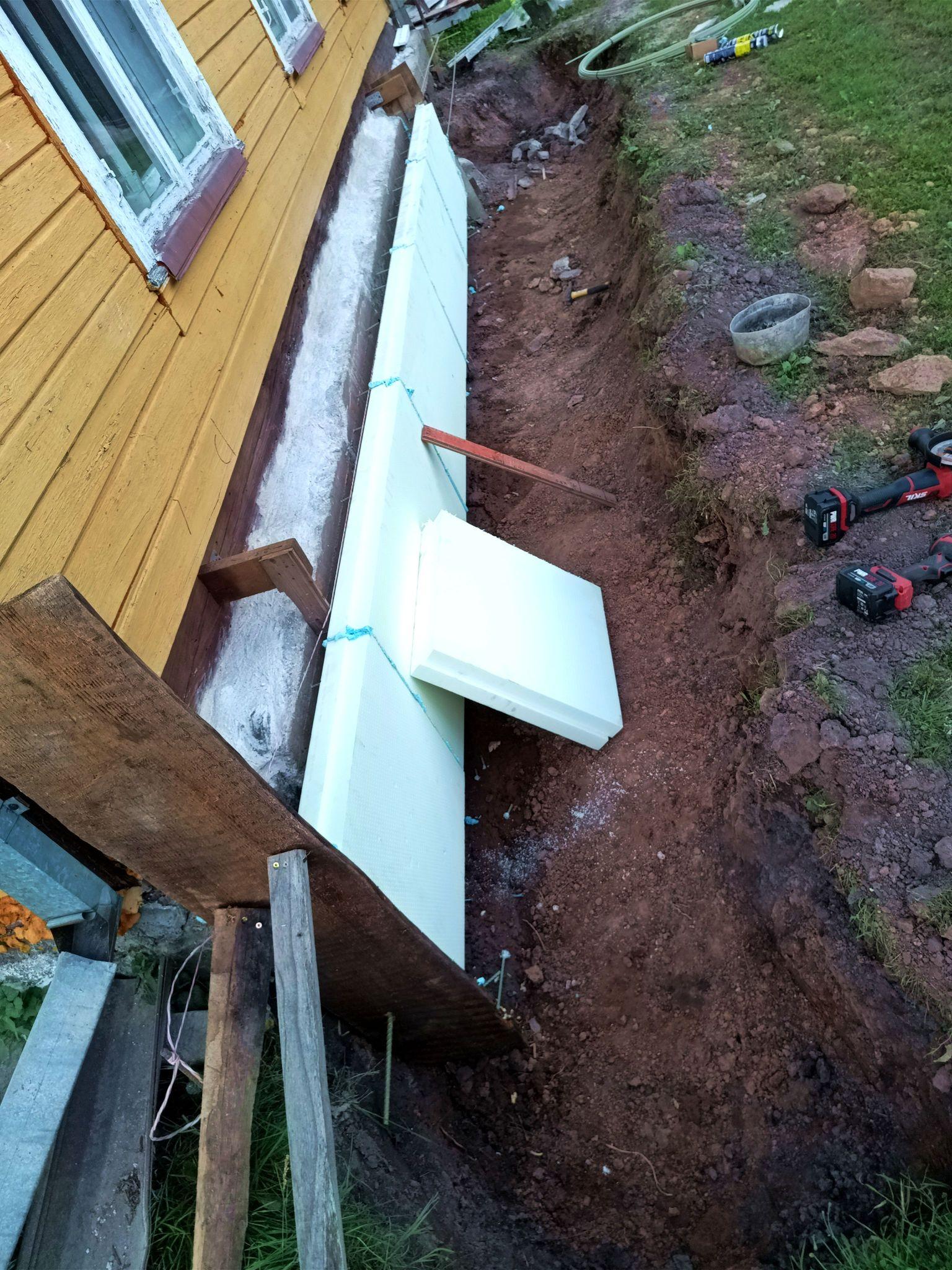 In fading light, a row of thick XPS foam panels is installed, hovering in the air above the ground. Blue glue binds them together and a plank and half a panel lean against them. The ends have also been closed with wooden boards that are supported with scrap wood pieces. Behind the foam some fibreglass rebar pieces are barely visible.