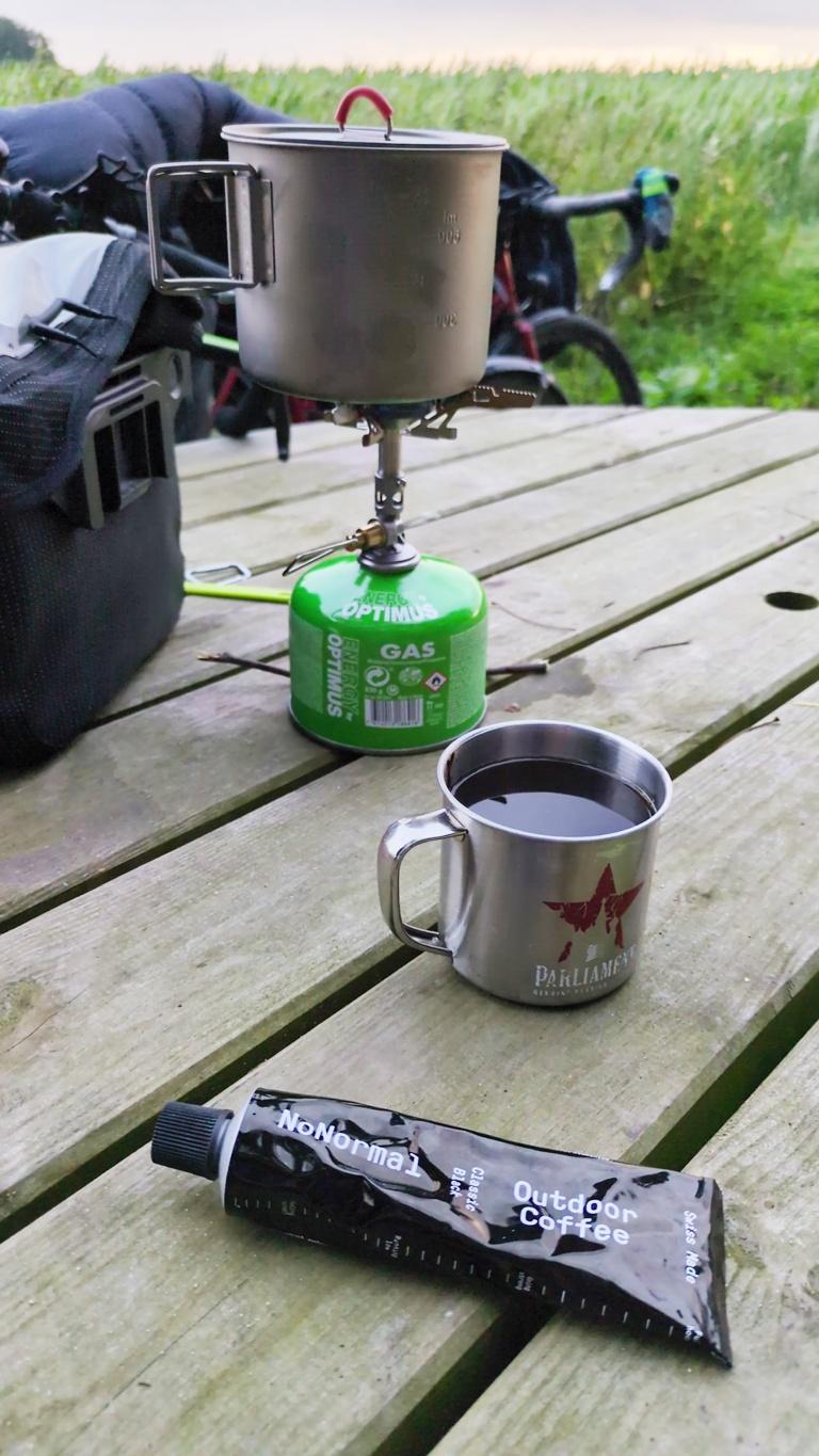 Eine schwarze Tube Outdoor Coffee, ein Metallbecher mit Kaffee und der Gaskocher mit Topf auf einem Holztisch.