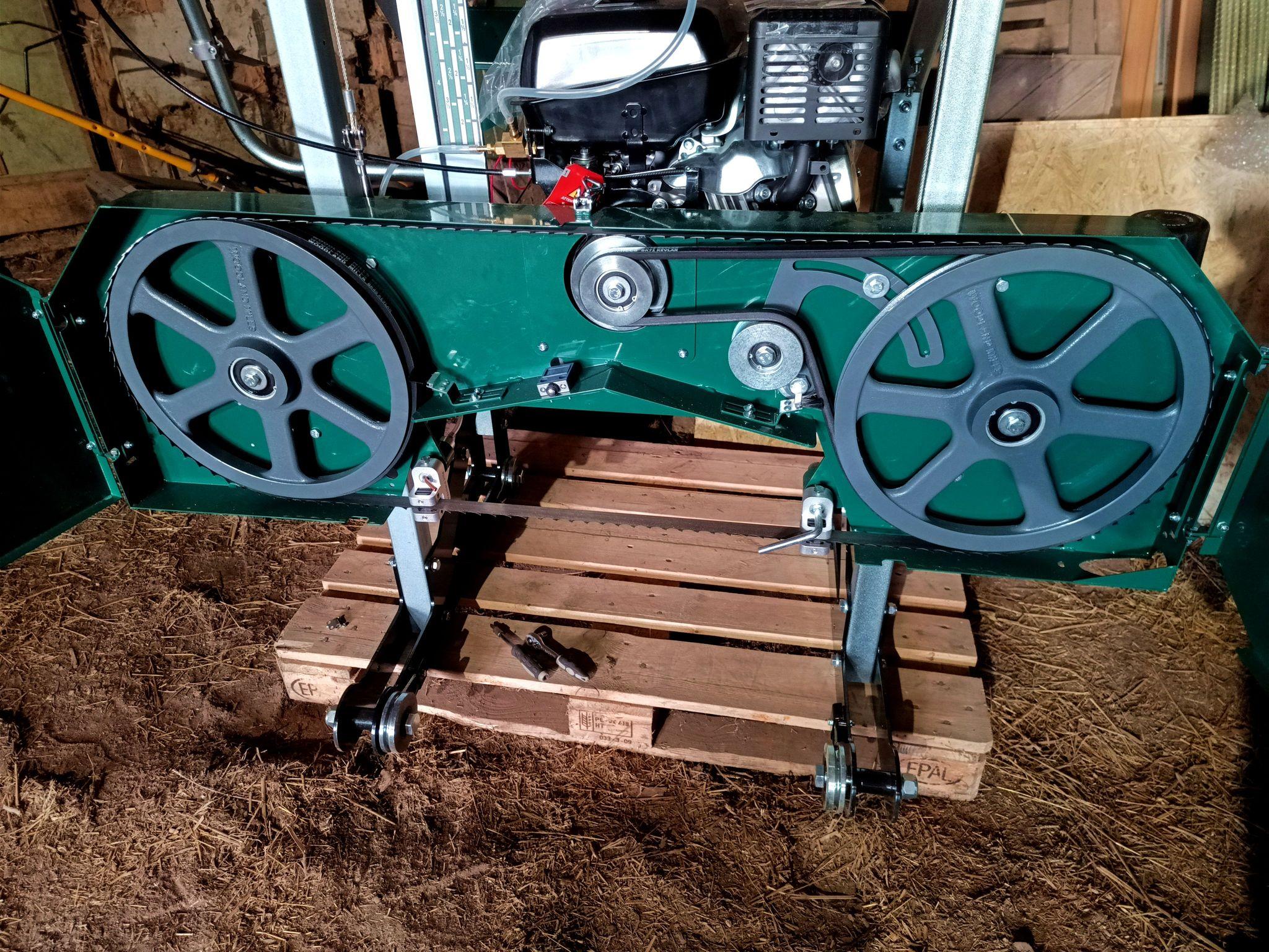 A bandsaw mill head sitting on a cargo pallet. The covers are open, exposing the drive wheel on the right and the idler wheel on the left, both with a belt on them. A sawblade is installed on them. There's a petrol engine on top of the lifting frame with a few stickers on it reminding to fill some oil in first.