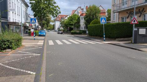 Man blickt ein breite, schnurgerade Straße hinunter. In der Mitte des Fotos quert ein Zebrastreifen die Fahrbahn und wird zusätzlich links und rechts von zwei großen Hinweistafeln angezeigt. Die Straße ist gesäumt von einigen kleinen Bäumen und Häuserzeilen auf beiden Seiten.