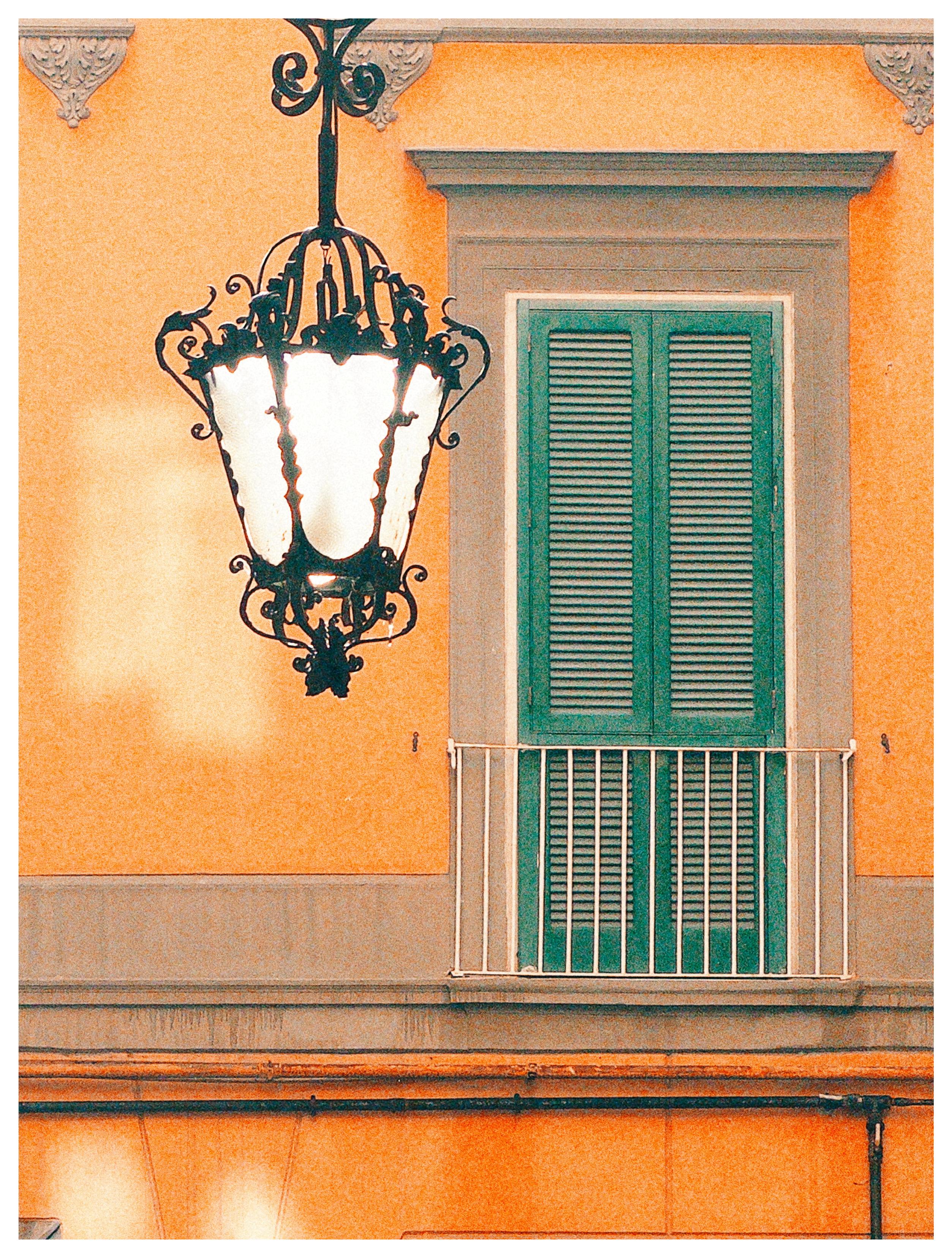 Ein großes Fenster mit grünen Fensterläden in einer orangenen Hauswand, davor noch eine Laterne