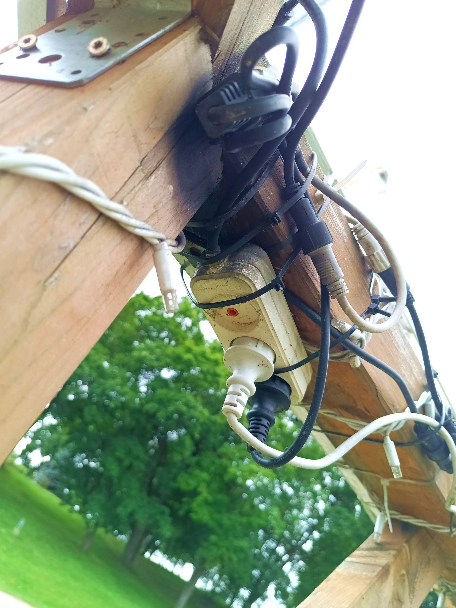 An indoor type triple electrical outlet, zip-tied to the bottom of a wooden beam, outside in a park by a lake. Its a GOST type, which looks like Schuko but without the PE contacts. The corner of the beam looks scorched and the side of the outlet is blackened.