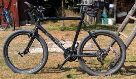 Ein sog. Urban-Bike lehnt mit dem Hinterrad an der Strebe eine Holzschaukel. Es handelt sich um ein schwarzes Scott Sub 30 mit nachgerüsteten hydraulischen Felgenbremsen. Der Rasen auf dem es steht ist leicht gelblich.