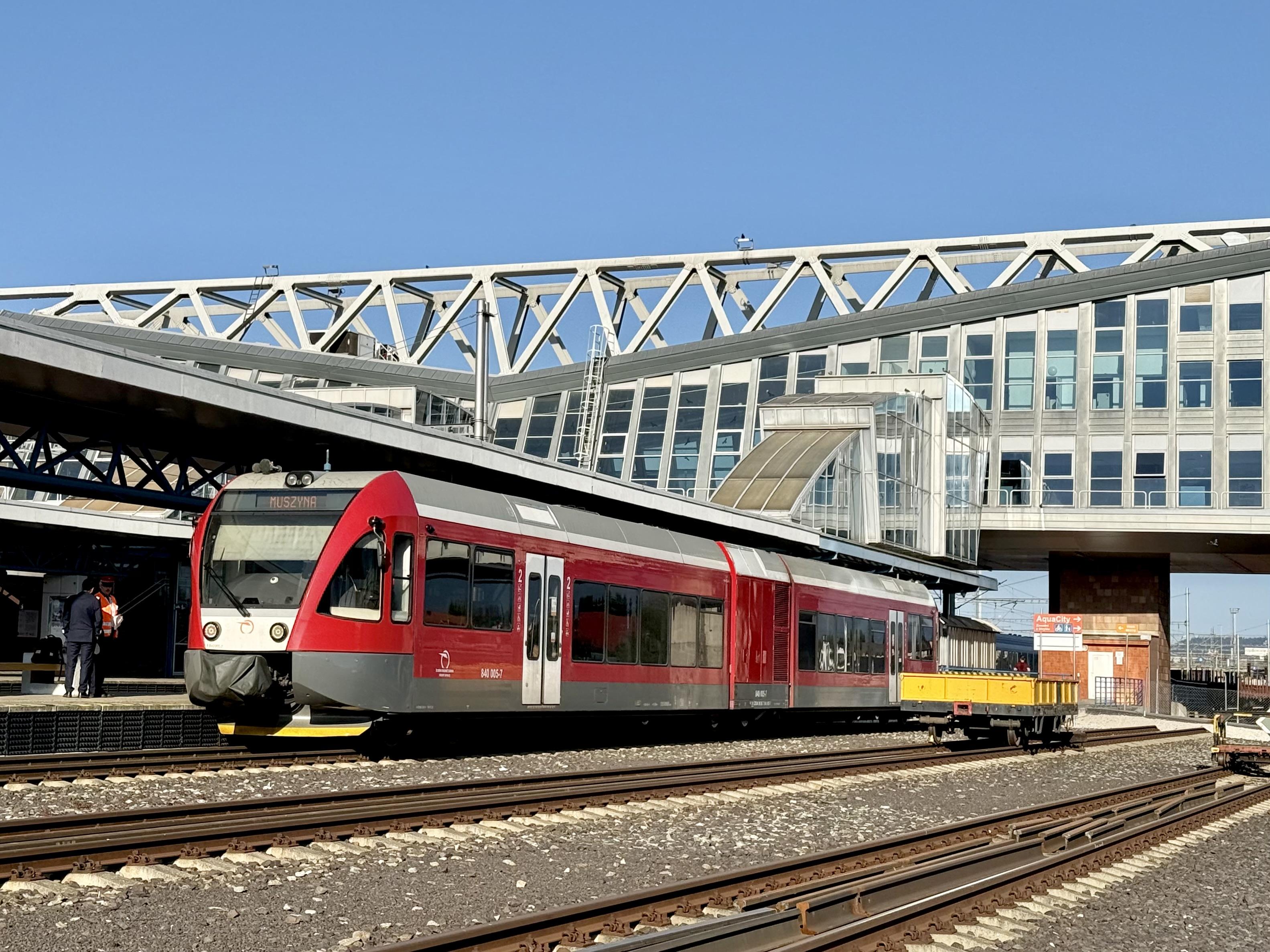 A red diesel train is waiting at a platform, photographed from beyond further station tracks with a steel and glass covered building bridging over the tracks in the background.