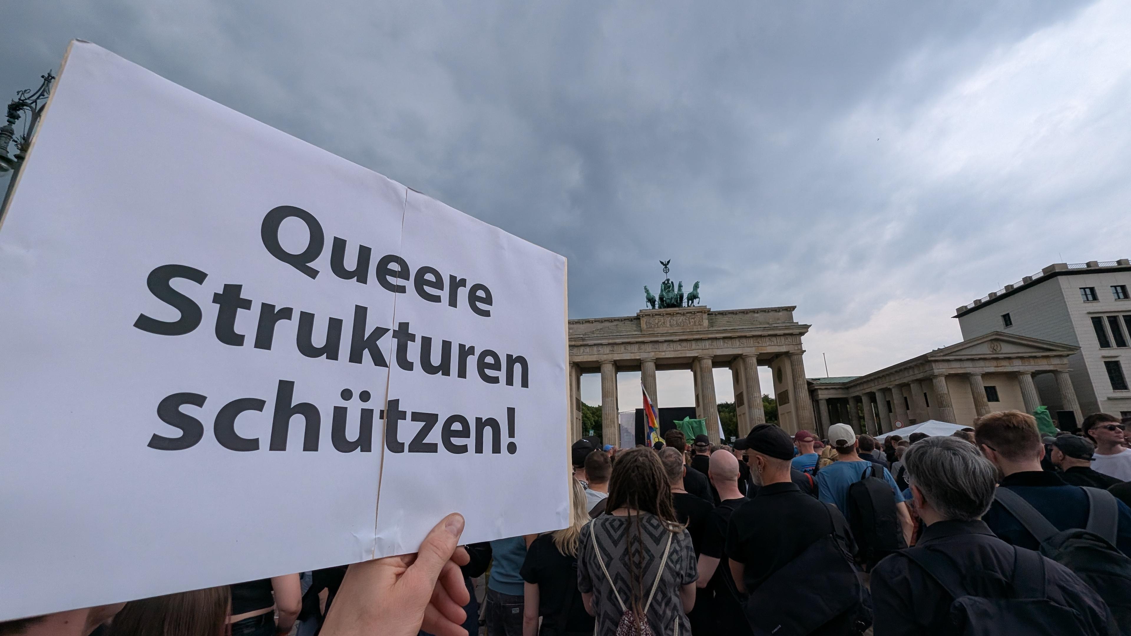 Plakat mit der Aufschrift Queere Strukturen schützen. Im Hintergrund ist das Brandenburger Tor