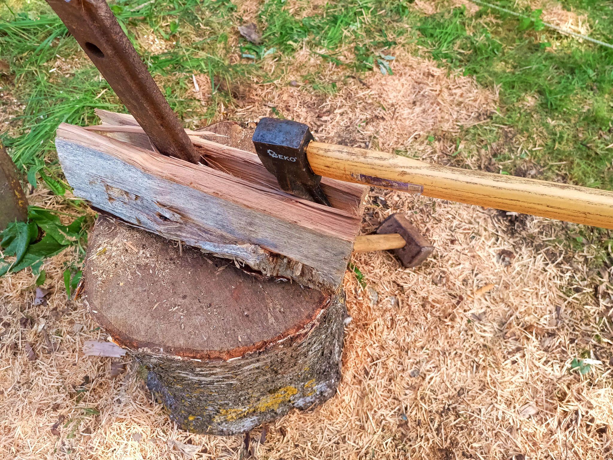 A birch log is being split on a chopping block with a large steel wedge and an axe stuck in a crack. Sawdust is piled up on the ground.