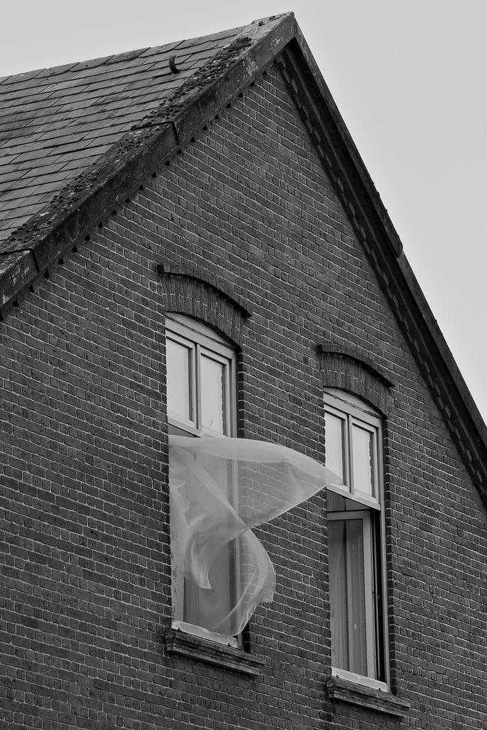 Zwei Dachgeschoßfenster an der Stirnseite eines Hauses. Schräg seitlich aufgenommen. Beide Fenster sind geöffnet, aus dem linken wehen die Gardinen wegen dem Durchzug raus.