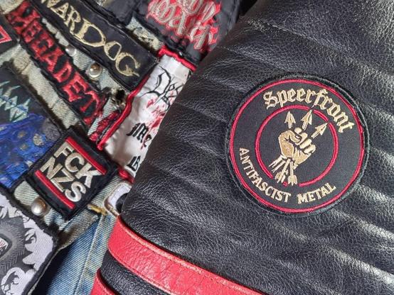 Speerfront-Patch auf Ärmel einer Lederjacke, daneben ist eine Stück Kutte mit FCK NZS Aufnäher zu sehen.