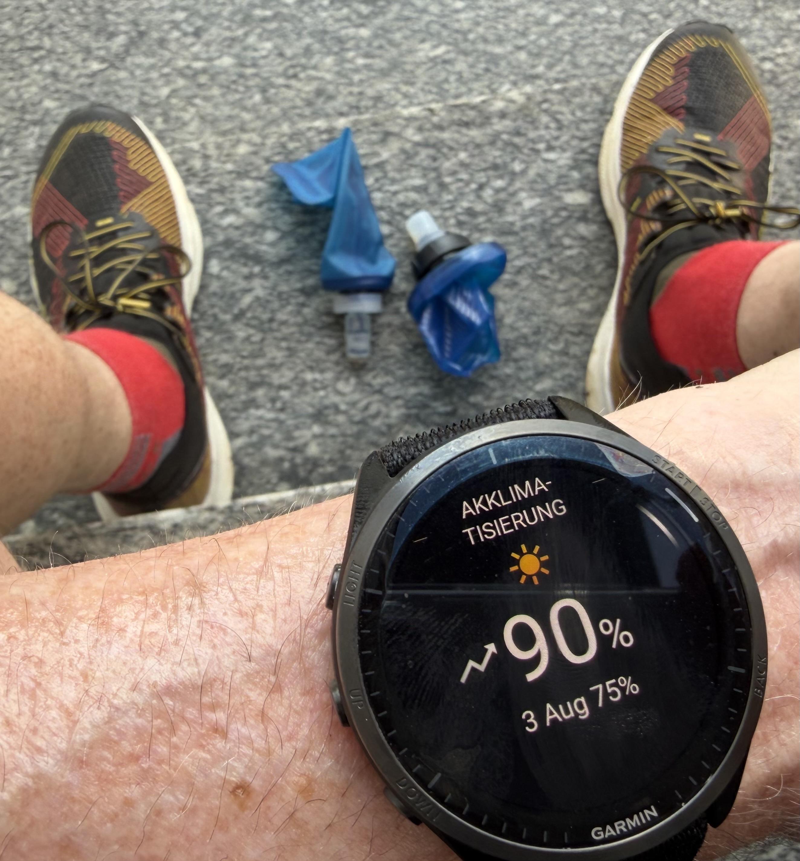 A close-up of a wrist showing a Garmin watch displaying a weather reading of 90% humidity, alongside a pair of colorful running shoes and two blue water bottles. The background features a textured surface.