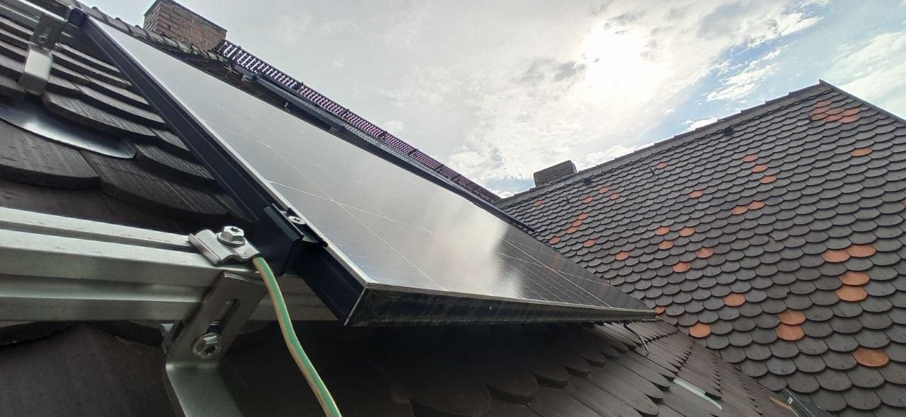 man sieht ein Stück altes Ziegeldach auf dem ein PV-Panel motiert ist. Ganz vorn im Bild ein gelb/grünes Erdungskabel und die Profilschienen zur Befestigung. Rechts im Bild der Teil vom Dach, der zum Giebel des Hauses führt und das PV-Modul am Vormittag verschattet.
