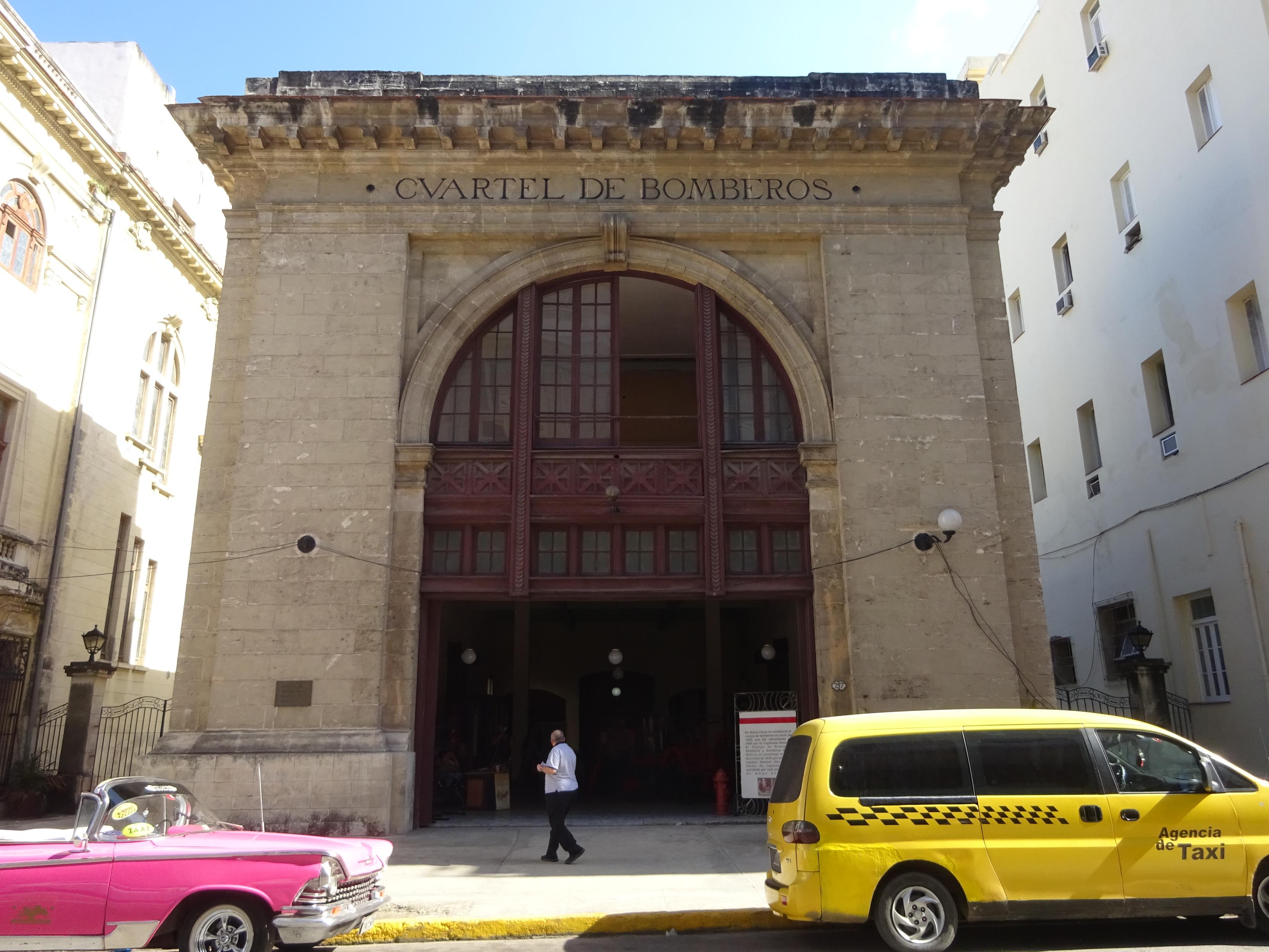 Ein historisches Gebäude mit einem großen Rundbogenportal und der Aufschrift 'CUARTEL DE BOMBEROS' über dem Eingang. Vor dem Gebäude stehen ein gelbes Taxi mit schwarzem Karomuster und ein pinkfarbenes Cabriolet. Eine Person in heller Kleidung geht auf den Eingang zu. Das Gebäude ist von weiteren hellen Fassaden flankiert, der Himmel ist blau mit einigen Wolkenteilen.