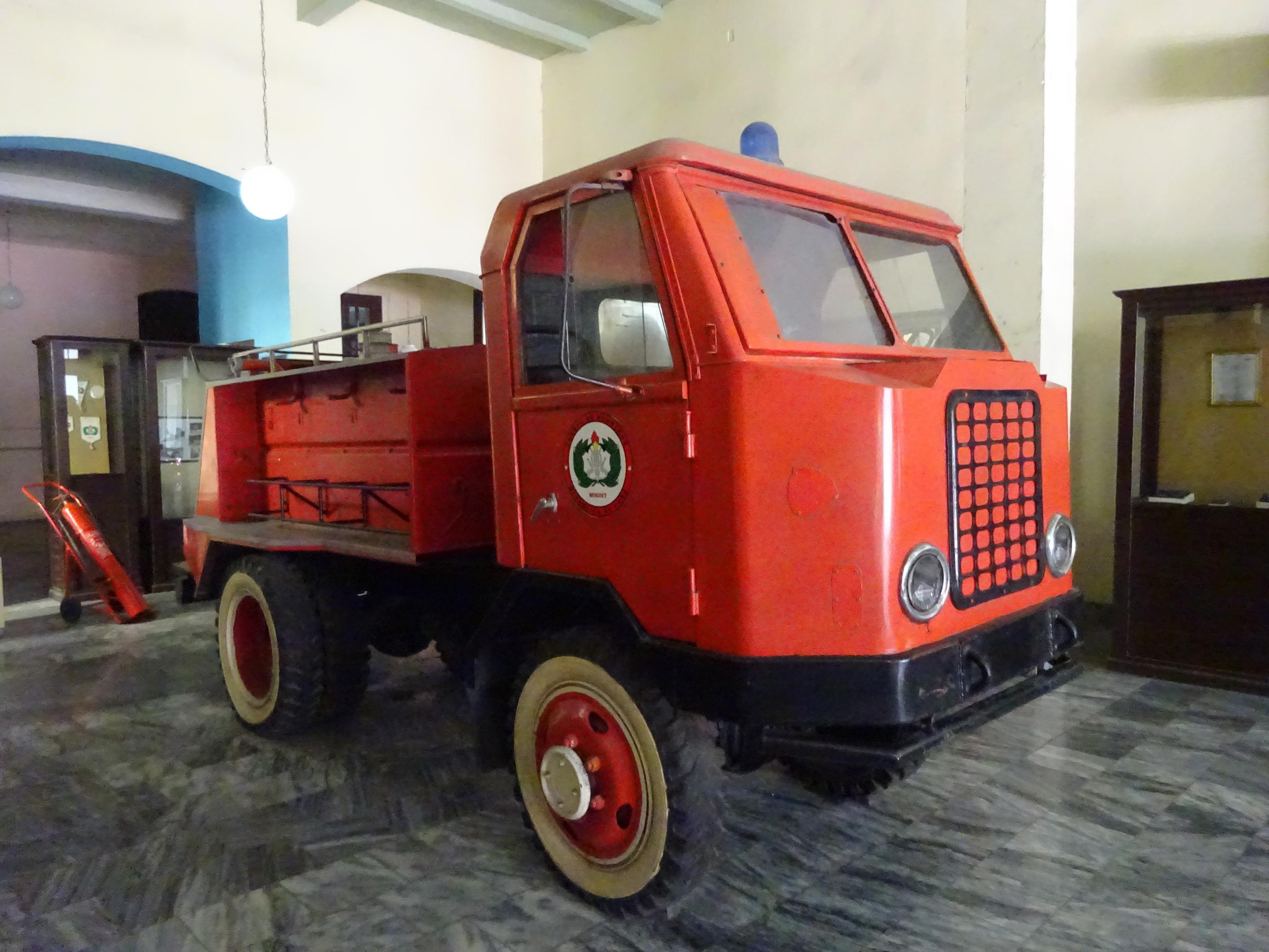 Ein roter Lastwagen mit eckiger Fahrerkabine steht auf einem Marmorboden in einem Innenraum. Die Front ist mit einem markanten schwarzen Gitter versehen. An der Seite befindet sich ein rundes Emblem mit grünen und weißen Elementen. Die Räder sind rot mit beigem Rand. Im Hintergrund sind dunkle Holzvitrinen und ein Handwagen sichtbar.