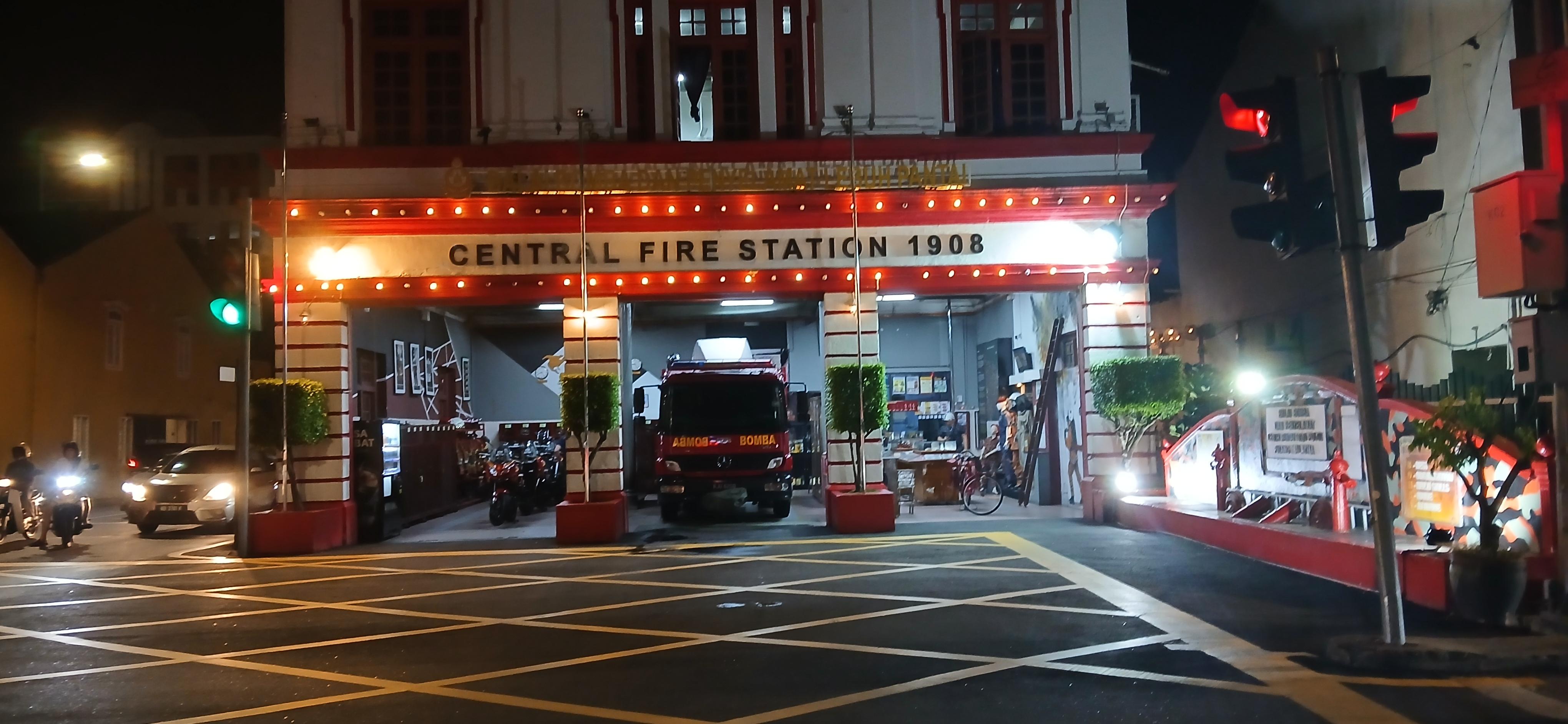 Im Mittelpunkt steht ein beleuchtetes Gebäude mit der Aufschrift 'CENTRAL FIRE STATION 1908' über dem Eingang. Ein rotes Feuerwehrfahrzeug ist vor dem Eingang geparkt. Die Szene spielt bei Nacht, die Straßenbeleuchtung und die Lichter am Gebäude sorgen für eine helle Atmosphäre. Vor dem Gebäude sind gelbe Markierungen auf der Fahrbahn zu sehen. Rechts im Bild befindet sich eine Ampel, die auf Rot steht. Links und rechts vom Eingang sind kleine, rechteckige Grünflächen mit niedrigen Bäumen angelegt.