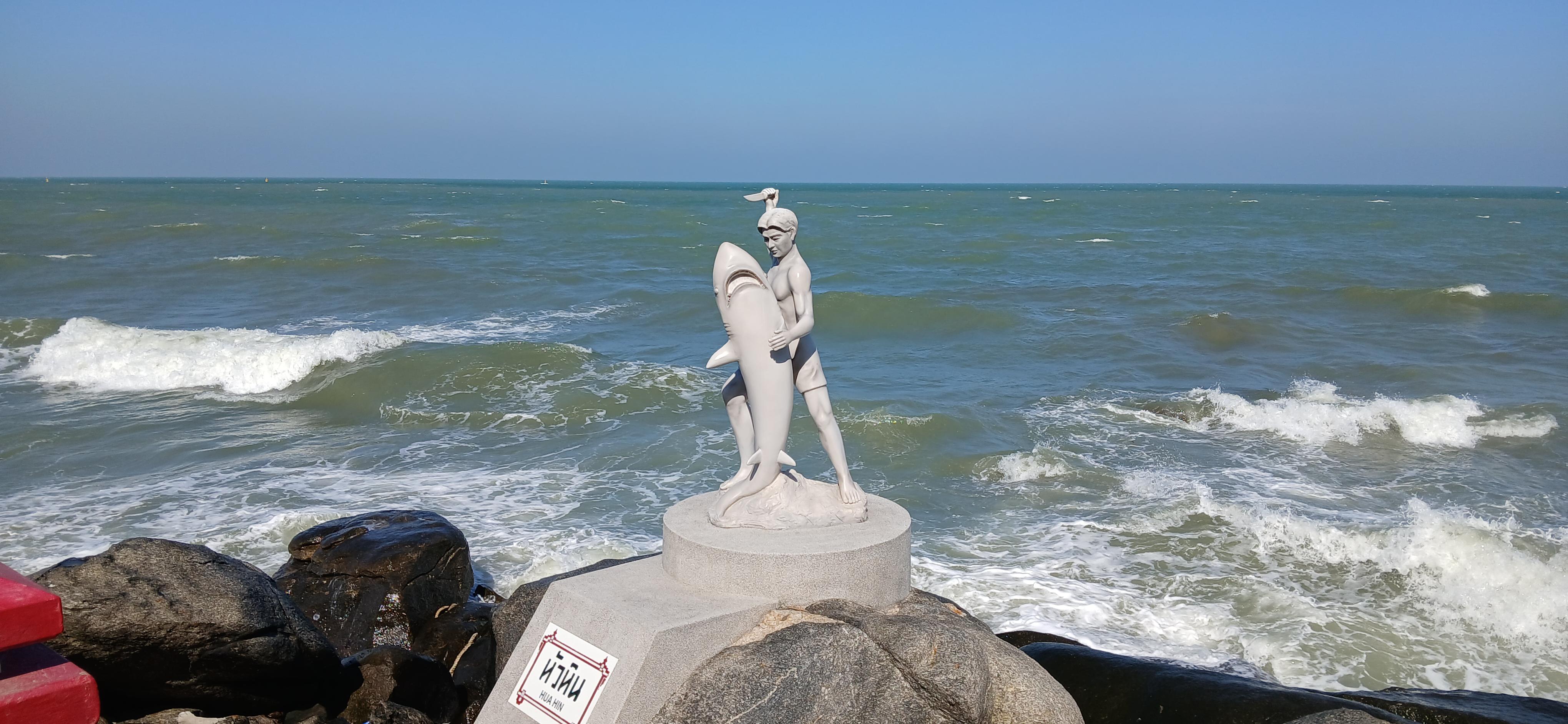 Im Vordergrund steht eine helle Statue auf einem runden Sockel, die eine Person mit einem großen Fisch/Hai zeigt. Die Figur trägt eine Kopfbedeckung und steht in dynamischer Pose. Im Hintergrund erstreckt sich das Meer mit weißen Wellen unter einem klaren blauen Himmel. Rechts und links sind dunkle Felsen zu sehen. Am Sockel befindet sich ein Schild mit kontrastreichem Text.