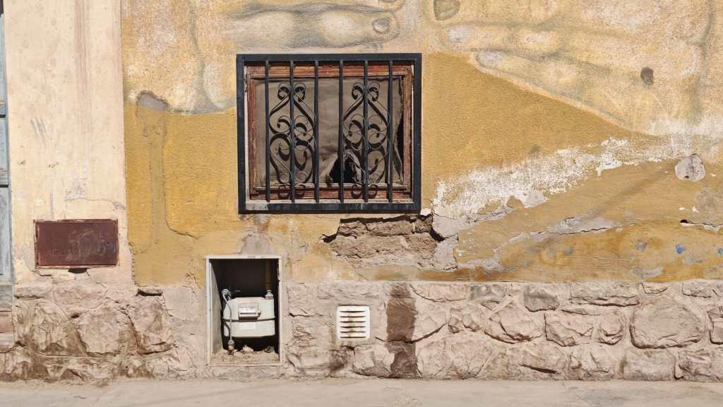 Vergittertes Fenster mit kaputten Fliegengitter. Der Putz bröckelt teilweise ab. 
Es gibt eine Gasuhr und Lüftungsgitter