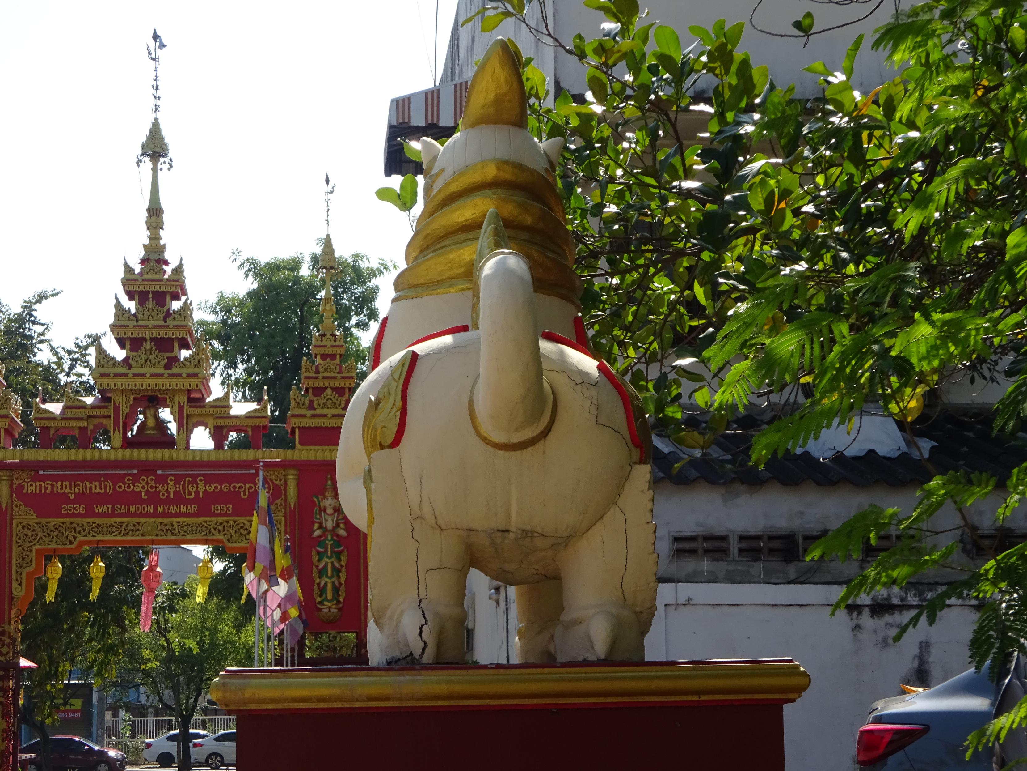 Im Vordergrund steht eine große, helle Statue eines Tieres mit goldfarbenen und roten Verzierungen auf einem Podest. Die Figur ist von hinten zu sehen und hat einen aufgerichteten Schwanz. Im Hintergrund befinden sich reich verzierte, rot-goldene Torbögen mit filigranen Details und goldenen Spitzen. Rechts ragen grüne Äste ins Bild, links sind bunte Fahnen zu erkennen. Die Szene spielt sich im Freien bei Tageslicht ab.