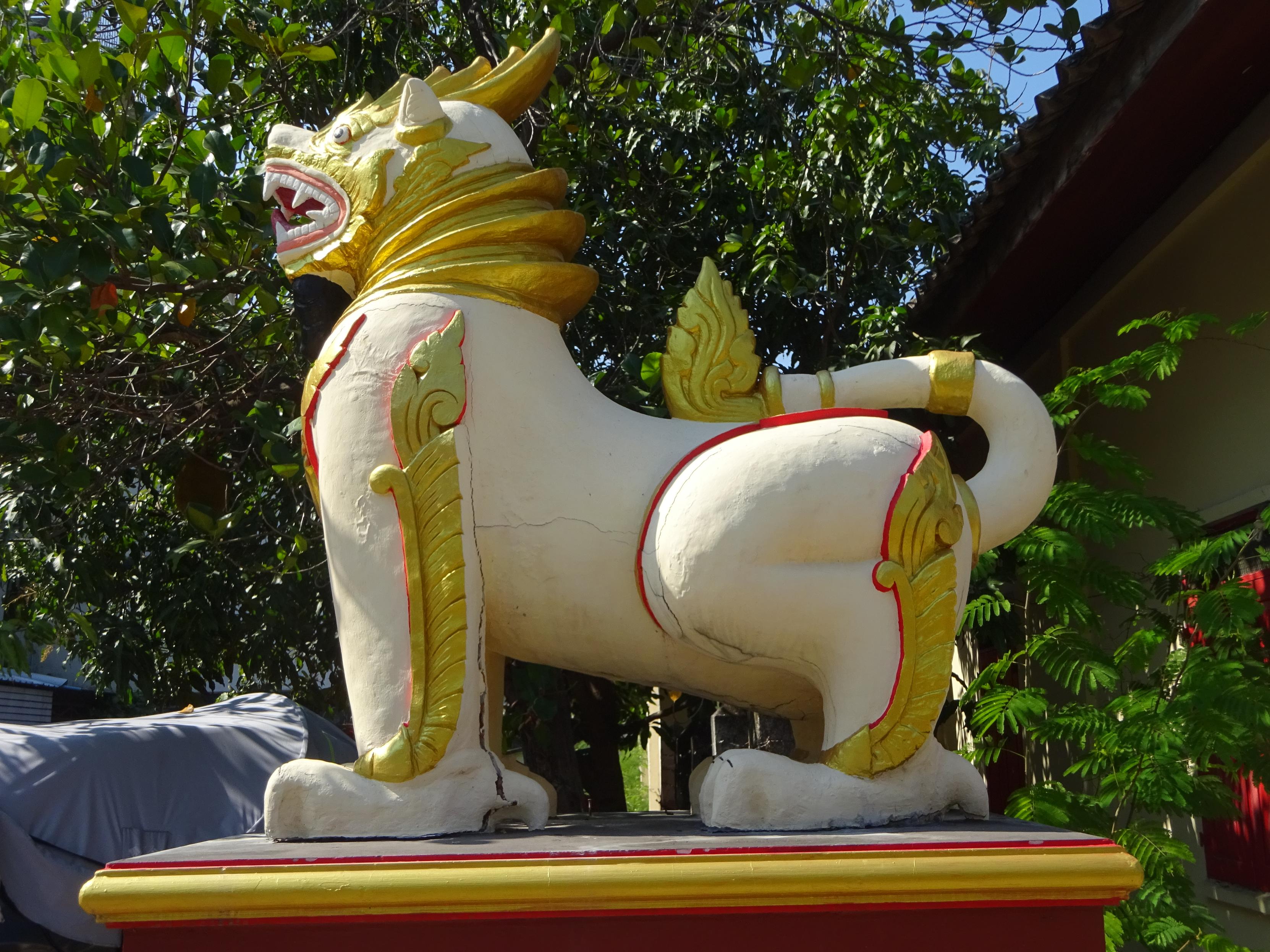 Eine große, stilisierte Löwenfigur steht auf einem roten Sockel. Die Skulptur ist überwiegend weiß und mit goldenen und roten Verzierungen versehen. Der Löwe hat einen geöffneten Mund mit sichtbaren Zähnen und eine goldene Mähne, die sich über den Rücken zieht. Im Hintergrund sind grüne Bäume und Teile eines Gebäudes zu sehen.