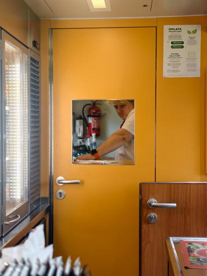 The actual kitchen on board where food is prepared; a photograph of the woman looking through a small window in the door, yellow interior 