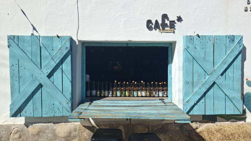 Ein offenes Fenster mit blauen Fensterläden. 
Auf der Fensterbank stehen viele Flaschen. 
Ein kleiner Klapptisch läd zum verweilen ein