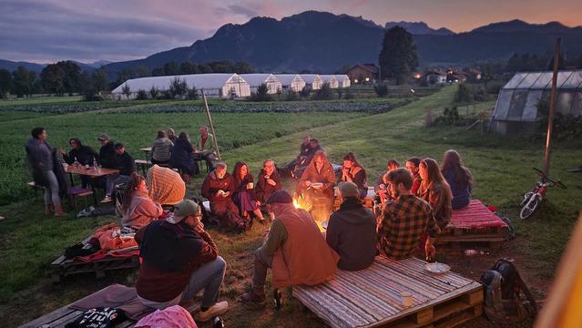 Foto von Menschen an einem Lagerfeuer, im Hintergrund Gewächshäuser und die Alpen mit Sonnenuntergang.