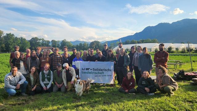 Foto von Menschen vor einem See auf einer Wiese, im Hintergrund die Alpen. In der Mitte ein Plakat des Netzwerk Solidarische Landwirtschaft e.V.